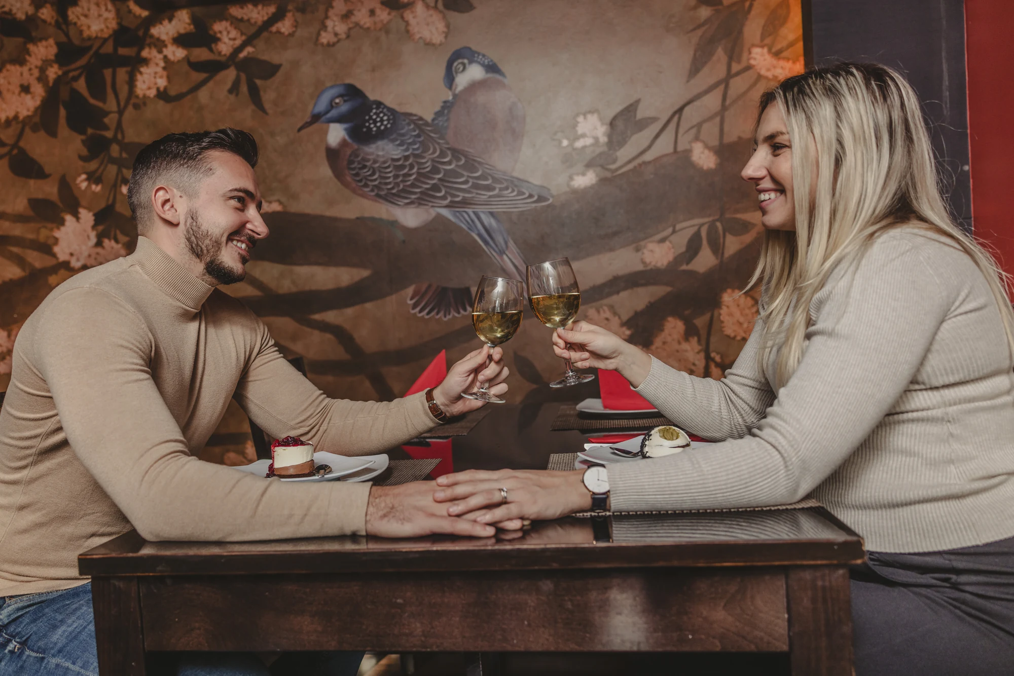 Cuplu care ciocnește pahare la o masă de restaurant, în fața unui perete ilustrat.