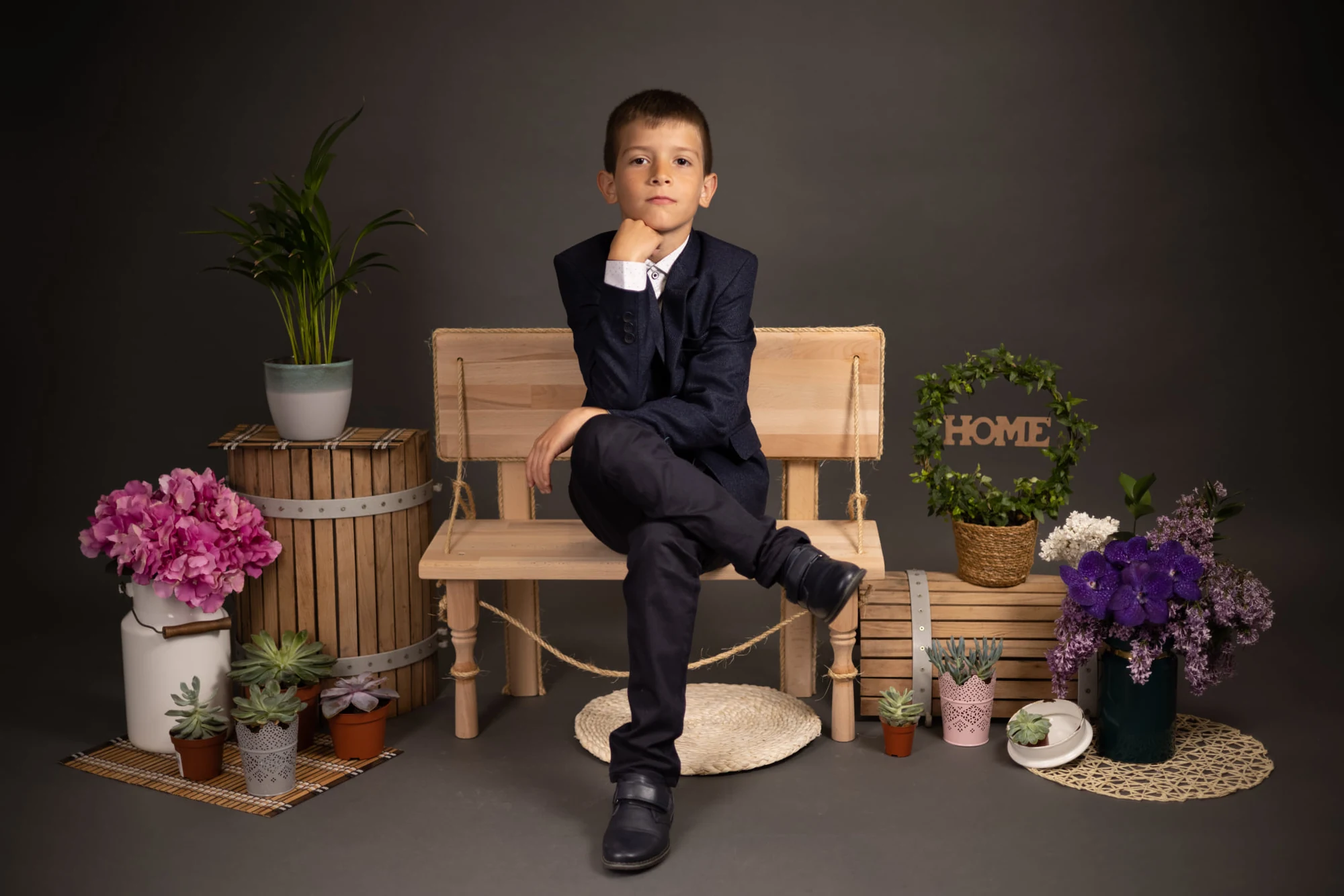Copil în costum așezat pe o bancă într-un decor de studio cu plante și flori.