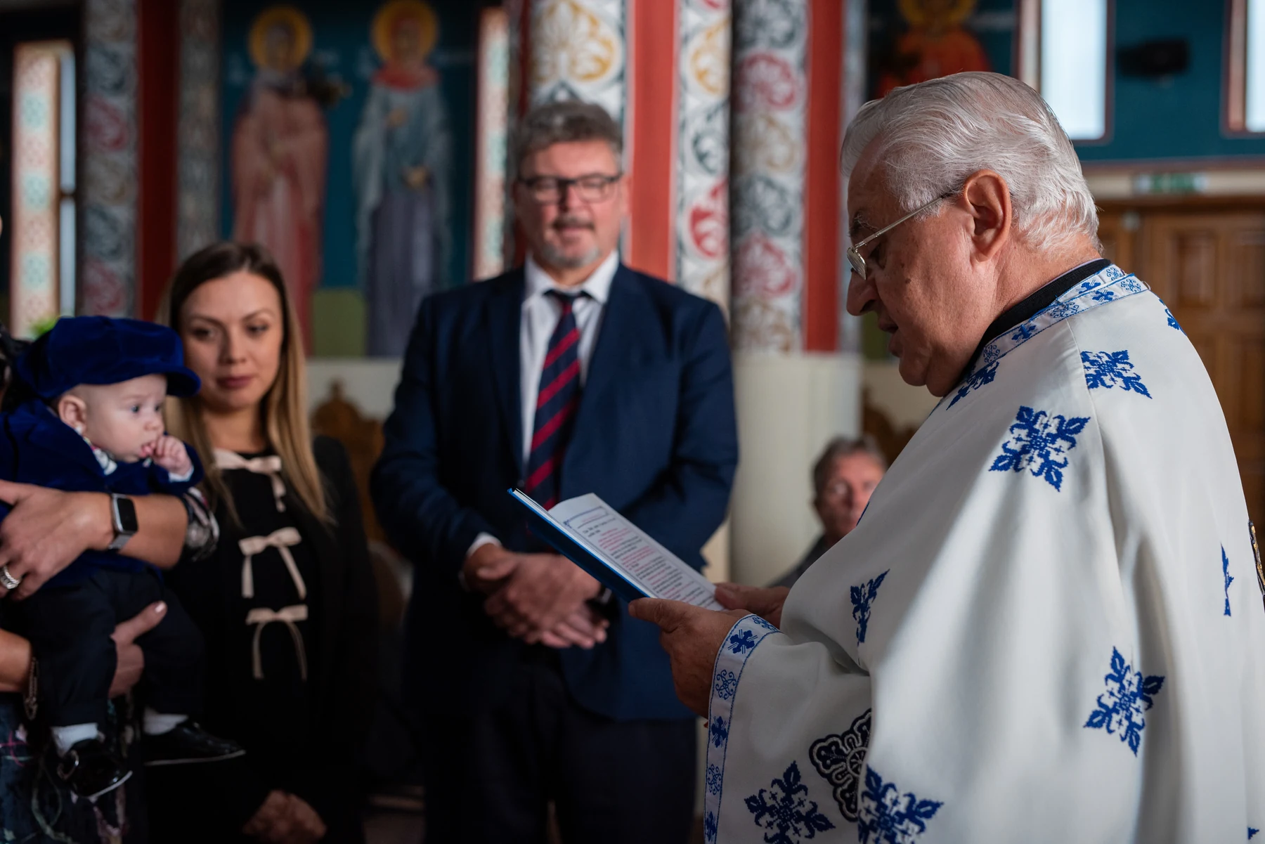 Preot citind în fața familiei și a bebelușului în timpul ceremoniei de botez.