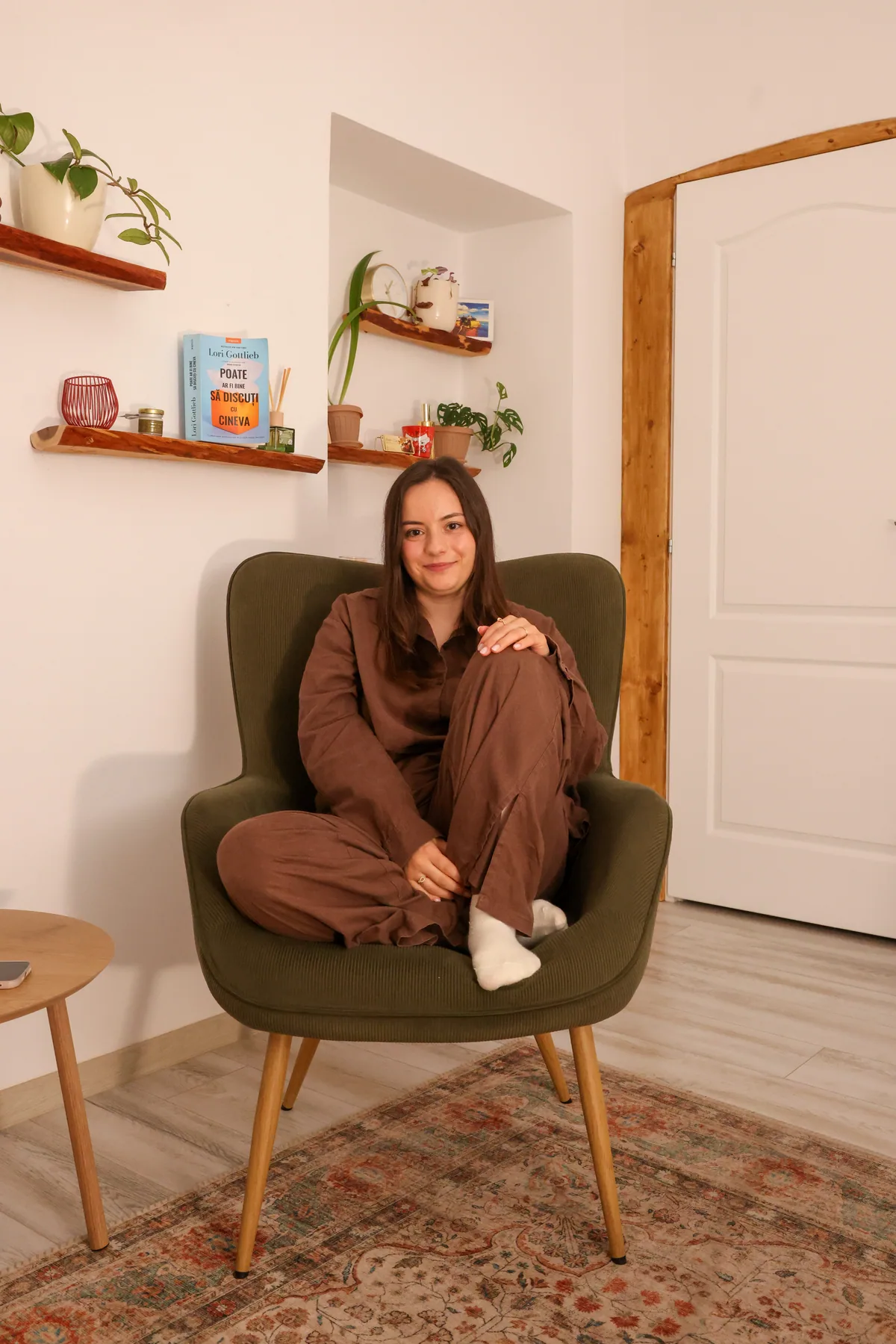 Femeie fotografiată într-un interior cald, așezată pe un fotoliu.